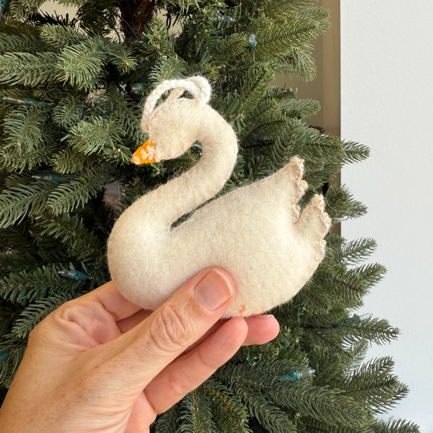 A hand holding a white felt swan ornament in front of a green Christmas tree.