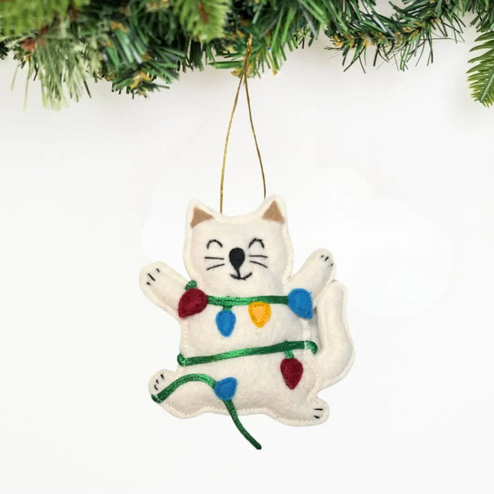 A white felt cat Christmas ornament hanging from a gold string, wrapped in green and multicolored thread with festive lights, set against a white background with pine branches at the top.