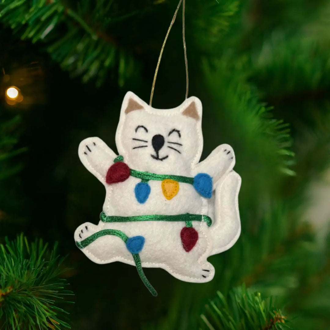 A white felt cat Christmas ornament with embroidered facial features and colorful string lights, hanging on a green Christmas tree branch.