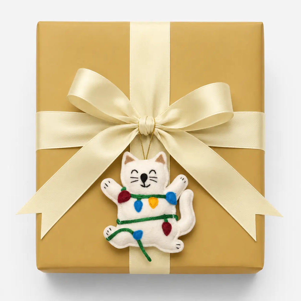 Gift box wrapped in gold paper with a white cat ornament holding Christmas lights on a white background