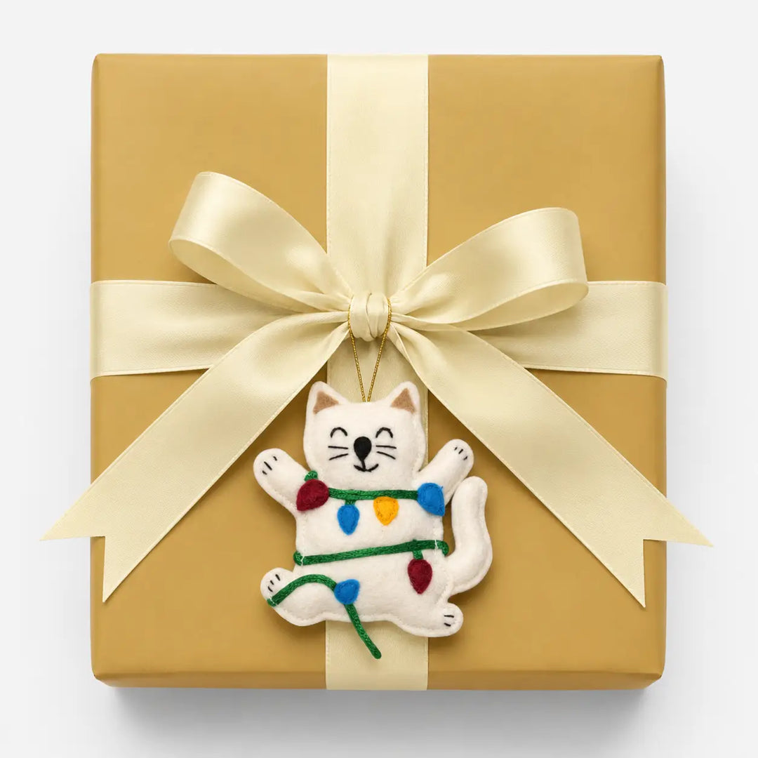 Gift box wrapped in gold paper with a white cat ornament holding Christmas lights on a white background