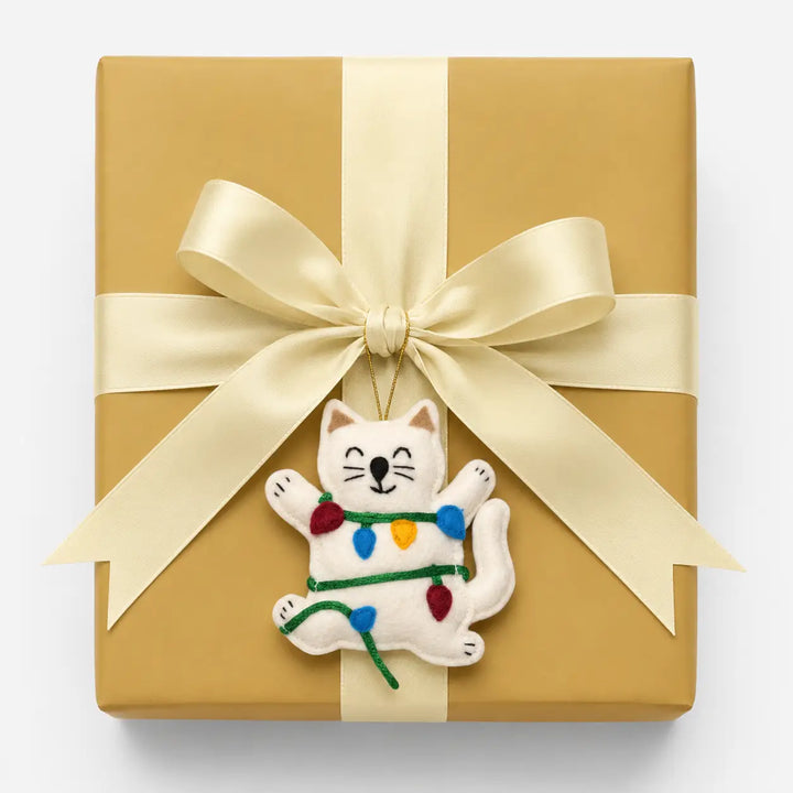 Gift box wrapped in gold paper with a white cat ornament holding Christmas lights on a white background