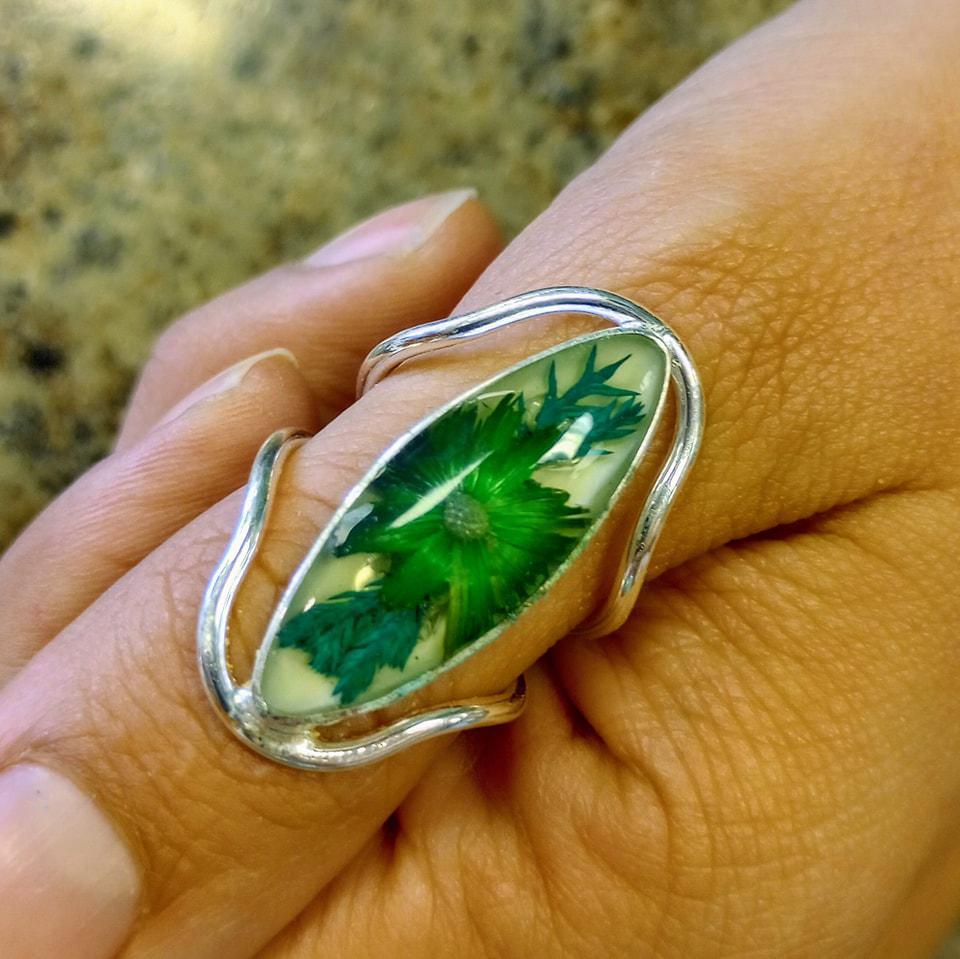 Silver ring with a resin center enclosing a flower and leaves, displayed on a hand.