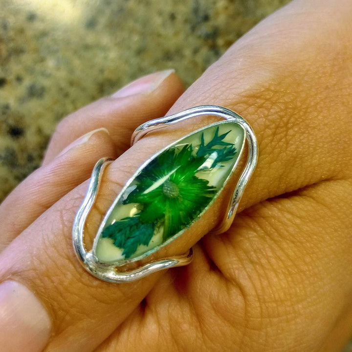 Silver ring with a resin center enclosing a flower and leaves, displayed on a hand.