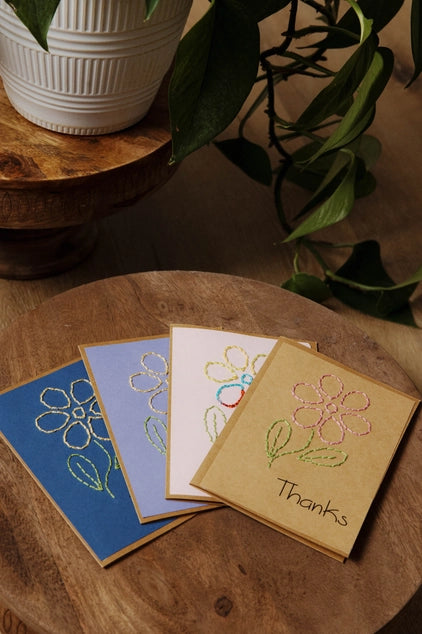 Four floral greeting cards on a wooden surface with plants in the background