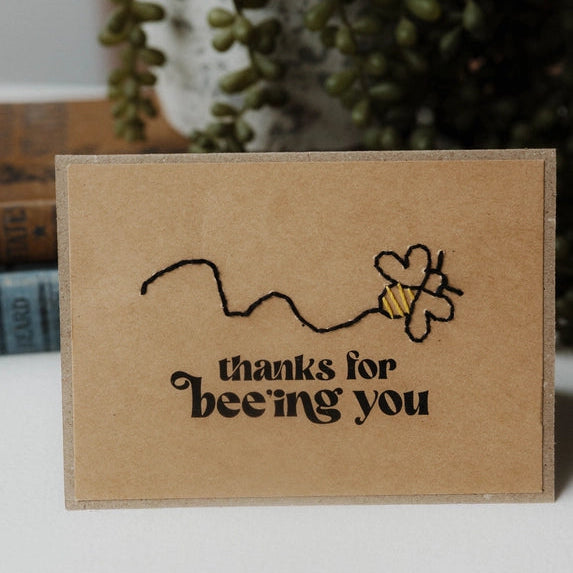 Kraft paper card with 'thanks for being you' text and a bee illustration, placed on a surface with a plant in the background.
