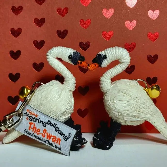 Two handmade string doll swans facing each other, made from white thread with black feet and orange beaks, on a red heart-patterned background. One swan has a keychain tag reading 'The Swan Handmade in Thailand' and gold bells.
