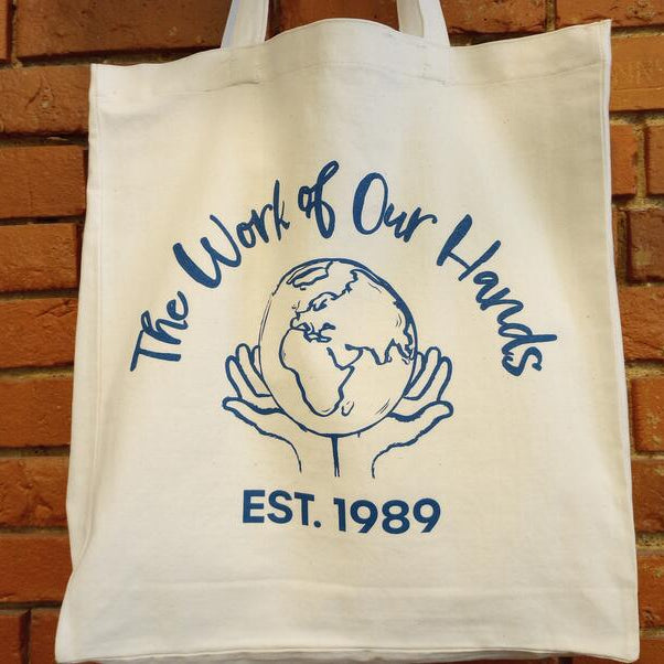 Tote bag with 'The Work of Our Hands' logo and text against a brick wall.