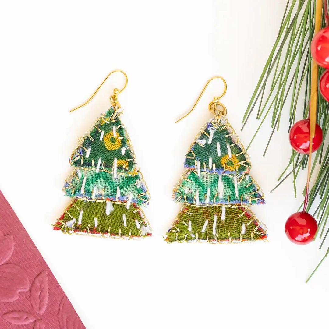A pair of handmade Christmas tree-shaped dangle earrings with gold fishhook wires and embroidered green, blue, and yellow tree bodies, displayed on a white background with pine branches and red berries as props.