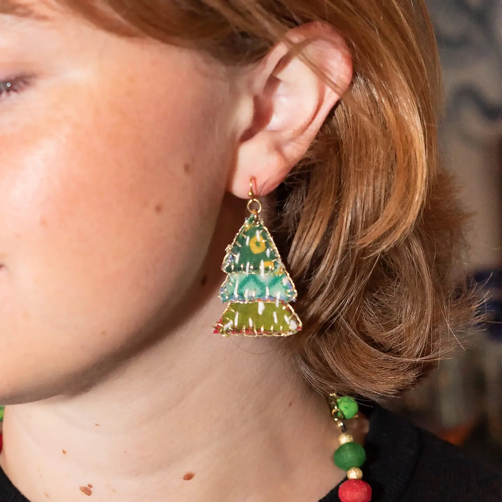 A close-up of a person wearing a dangling tree-shaped earring with a green and yellow fabric and gold-tone metal trim.