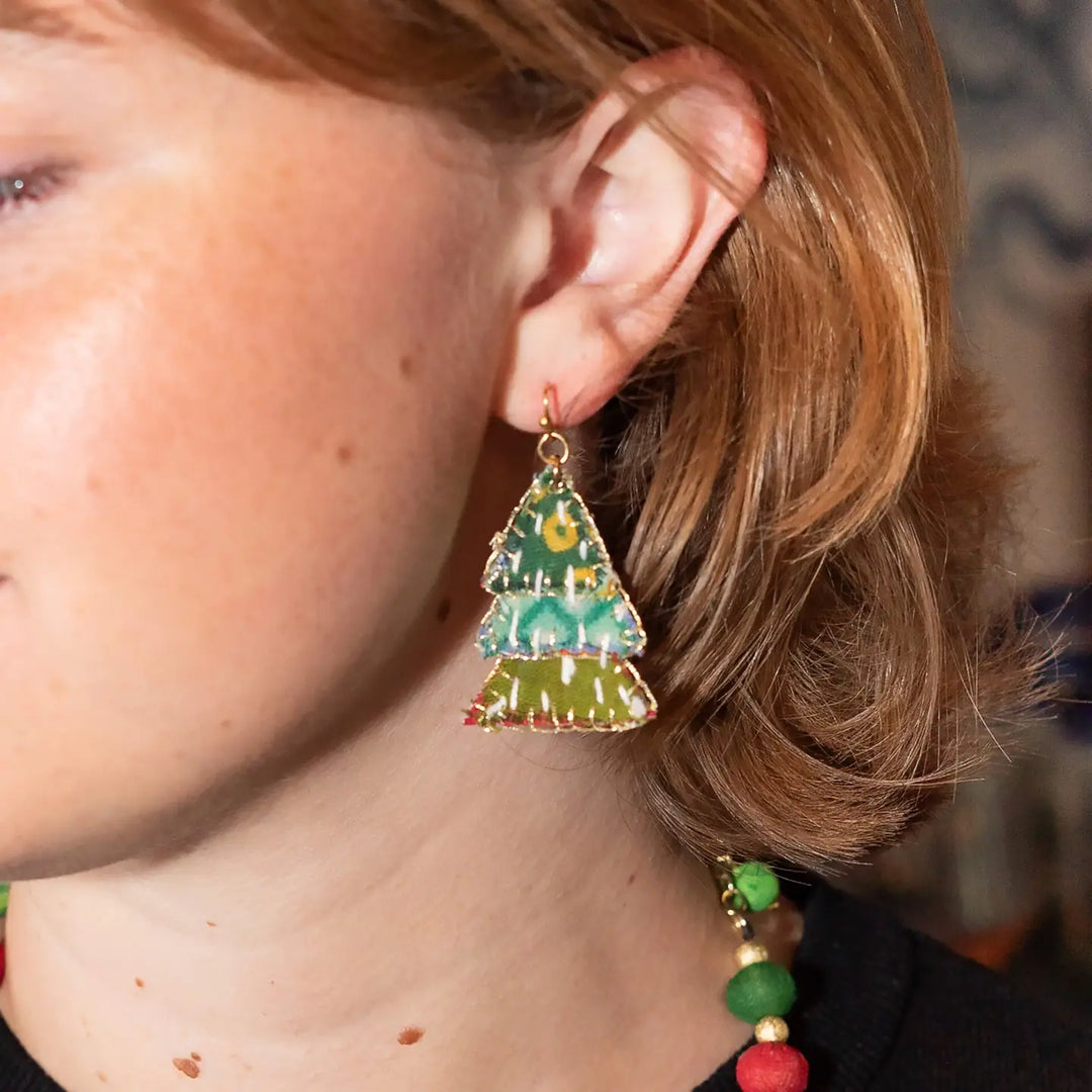 A close-up of a person wearing a dangling tree-shaped earring with a green and yellow fabric and gold-tone metal trim.