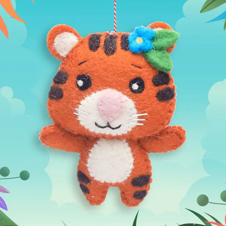 A cute orange felt tiger ornament with black stripes, a pink nose, white paws and face details, and a blue flower with a green leaf on its head, hanging against a light blue background with subtle decorative leaves.