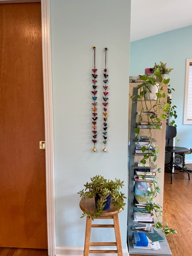 A pair of colorful hanging decorations displayed on a light blue wall in a home interior, with a wooden stool holding a potted plant below and a shelving unit with books and trailing green plants to the right.