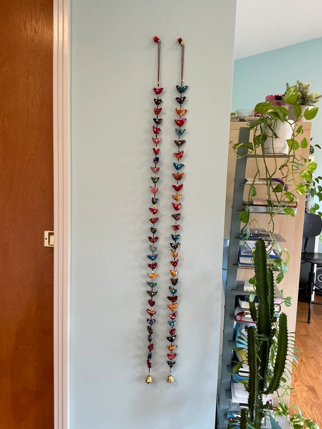 Two long vertical strands of colorful decorations hang on a light blue wall in a home interior, with a wooden door on the left and a shelving unit with plants on the right.