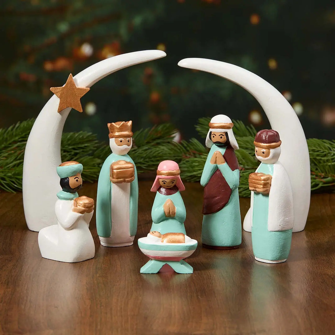 A wooden nativity scene figurine set on a dark wooden table with evergreen branches in the background. The set includes a crescent moon with a star, three wise men, Mary, and baby Jesus in hand-painted pastel colors.