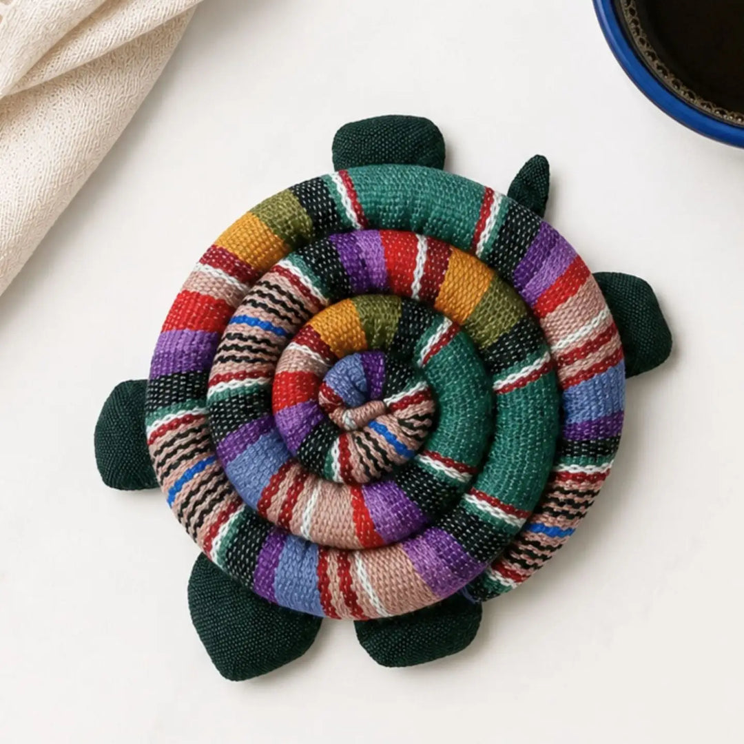 A handmade, spiral-shaped turtle-shaped trivet made from colorful woven textile strips, displayed on a light off-white surface with a beige fabric napkin and a dark blue cup partially visible.