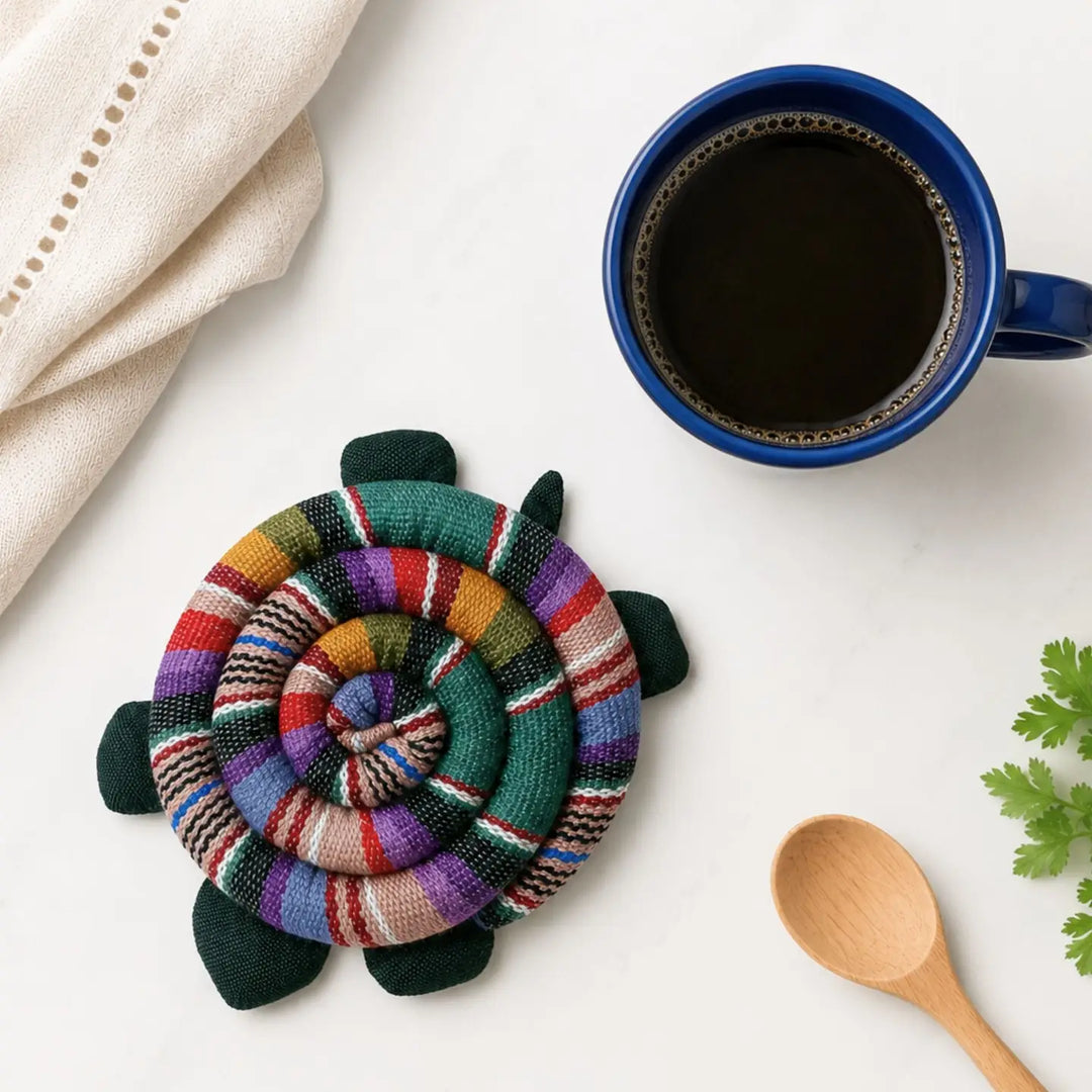 Colorful woven turtle  trivet on a white surface with a blue mug of coffee and wooden spoon.