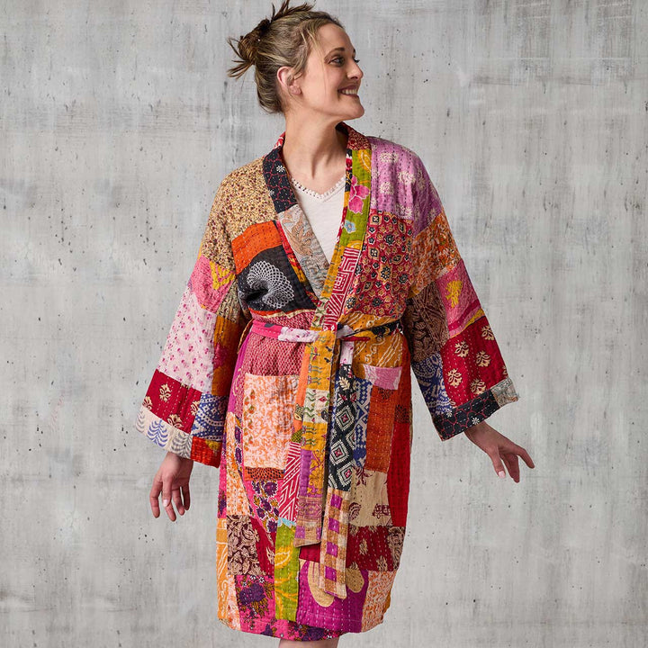 Woman wearing a colorful patchwork robe against a concrete wall.