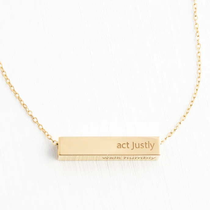 Gold necklace with a bar pendant engraved 'act Justly' on a white background
