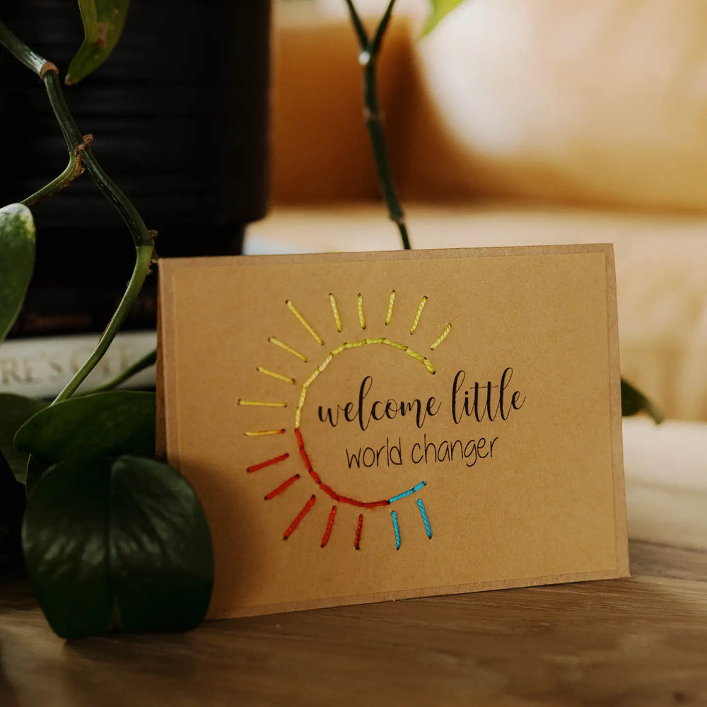 Brown greeting card with sun design and text 'welcome little world changer' on a wooden surface with plants.