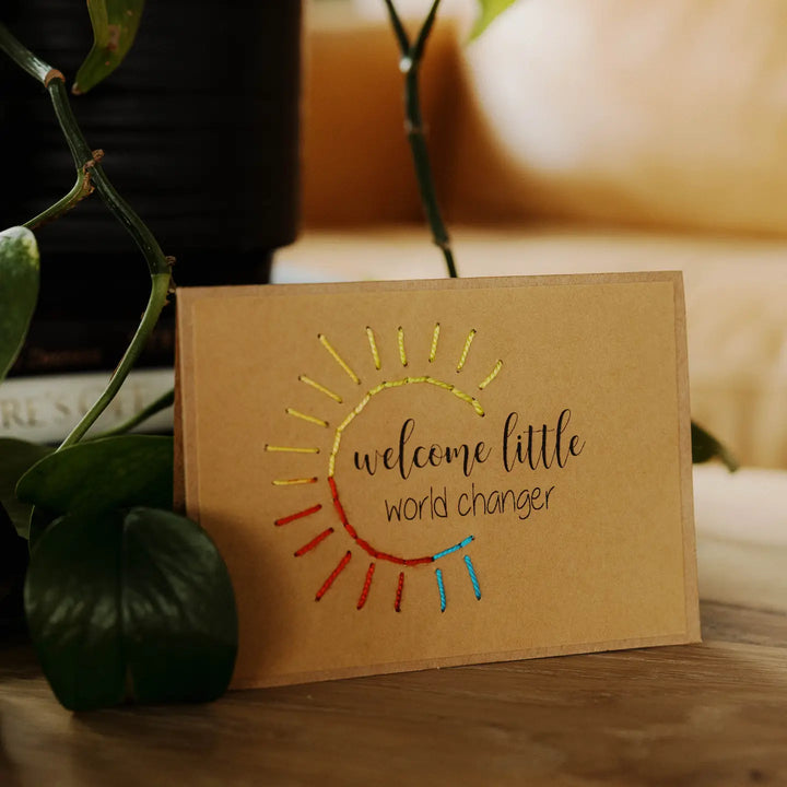 Brown greeting card with sun design and text 'welcome little world changer' on a wooden surface with plants.