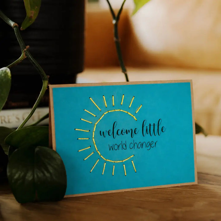 Blue card with yellow sun design and text 'welcome little world changer' on a wooden surface with plants.