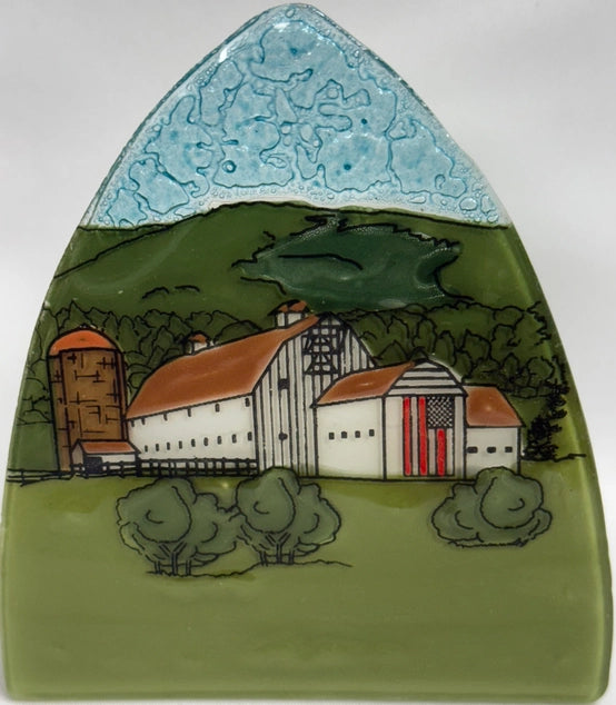 A hand-painted ceramic wall plaque depicting a farm scene with a white barn, silo, red roof, green hills, and a blue sky, set against a neutral gray background.