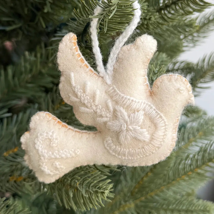 A cream-colored felt Christmas ornament shaped like a dove, hanging on a green pine tree branch.