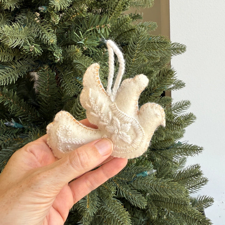 A hand holding a cream-colored felt Christmas ornament shaped like a dove in front of a green Christmas tree.