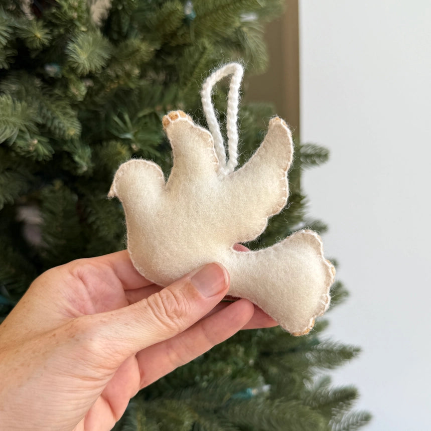 A hand holding a small felt Christmas ornament shaped like a dove in cream wool, with a green Christmas tree in the background.