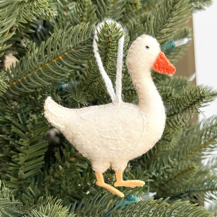 A felt Christmas ornament shaped like a white goose with an orange beak and yellow feet, hanging on a green pine tree branch.