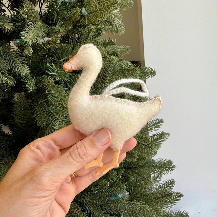 A hand holding a small felt goose ornament in front of a green Christmas tree and a light wall.