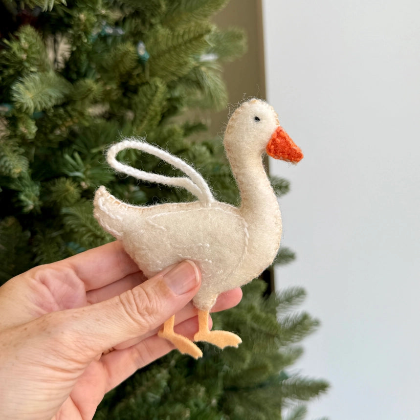 A hand holding a small felt goose ornament in cream with a bright orange beak and legs, with a green Christmas tree in the background.