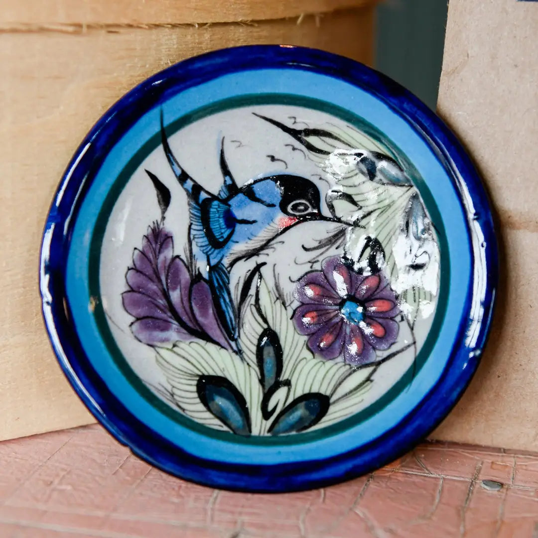 A round hand-painted ceramic plate with a cobalt blue rim and a central illustration of a hummingbird, purple flowers, and green leaves on a white background, leaning against a cardboard box.