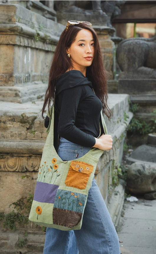 A woman wearing a black off the shoulder top and blue jeans stands outdoors in front of stone ruins, carrying a large patchwork fabric tote bag with floral and geometric embroidered panels.