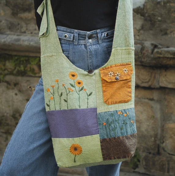 A person wearing a black top and blue jeans carries a large patchwork fabric shoulder bag with embroidered flowers and colored fabric panels, standing outdoors against a stone wall.