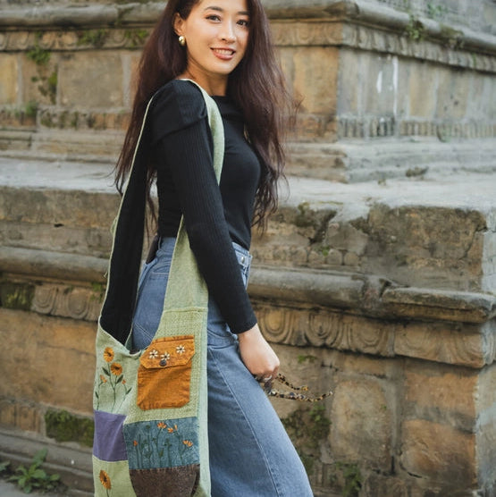 A woman wearing a black long-sleeve top and wide-leg blue jeans carries a large, colorful patchwork fabric shoulder bag with a green base and orange, yellow, and purple stitched details, standing in front of a stone staircase.