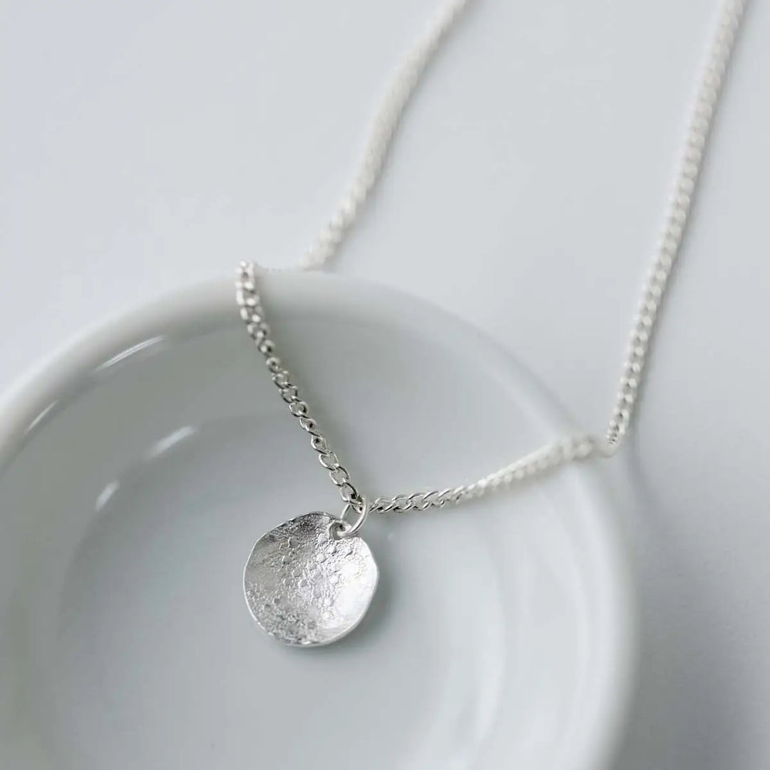 A silver pendant necklace with a textured, hammered circular disc charm on a delicate chain, displayed on a white ceramic dish against a light gray background.