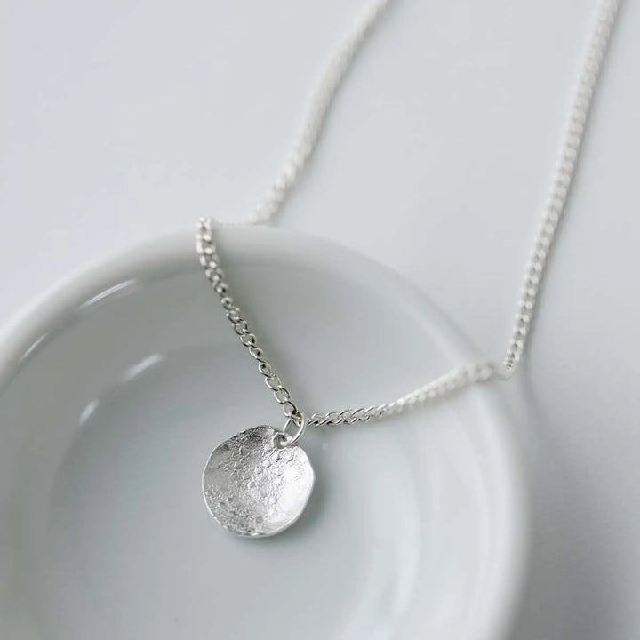 A silver pendant necklace with a textured, hammered circular disc charm on a delicate chain, displayed on a white ceramic dish against a light gray background.