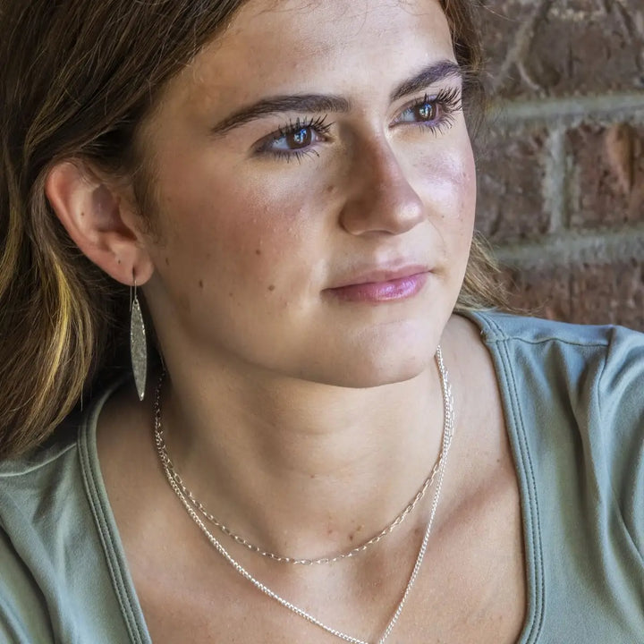 A close-up portrait of a woman wearing a silver dangle earring, layered silver necklaces, and a sage green top, set against a blurred brick wall background.