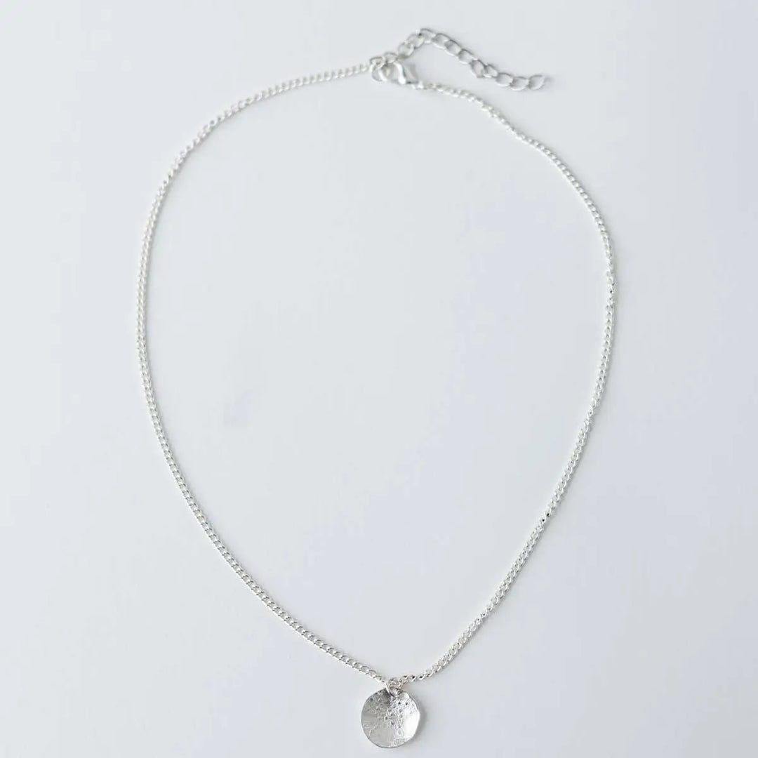 A delicate silver chain necklace with a small, textured circular disc pendant, displayed on a clean white background.