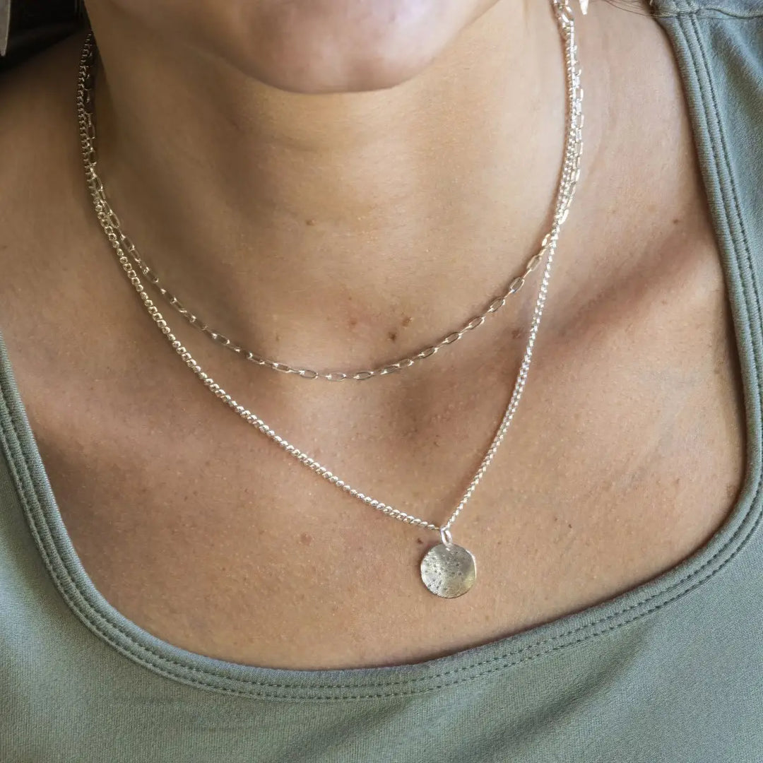 A close-up of a person's neck and chest wearing a layered necklace with a textured silver disc pendant on a fine chain, over an olive green top.