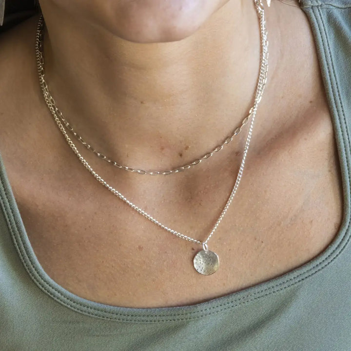 A close-up of a person's neck and chest wearing a layered necklace with a textured silver disc pendant on a fine chain, over an olive green top.