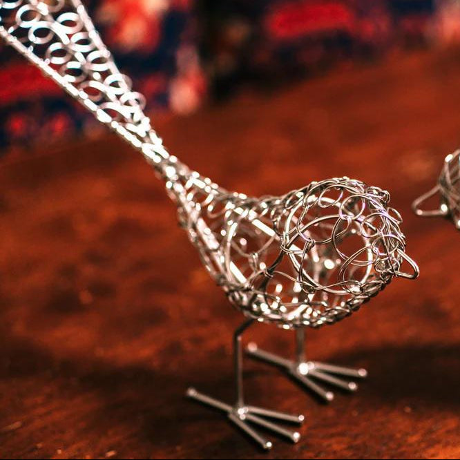 A silver wire sculpture of a bird displayed on a warm brown wooden surface with a blurred background.