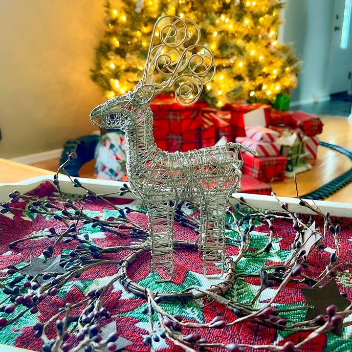 A silver wire reindeer with gold antlers stands on a red and green holiday table runner with berry branches, with a decorated Christmas tree and wrapped gifts blurred in the background.