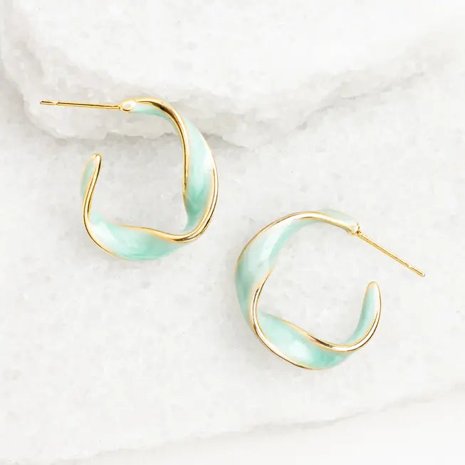 A pair of gold hoop earrings with a wavy, twisted design and a light mint green enamel inlay, displayed on a white textured stone surface.