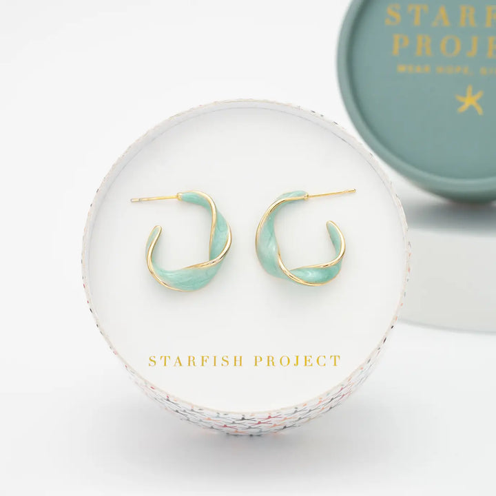 A pair of gold crescent hoop earrings with a twisted mint green enamel center, presented on a white round display card with the brand name 'STARFISH PROJECT'.
