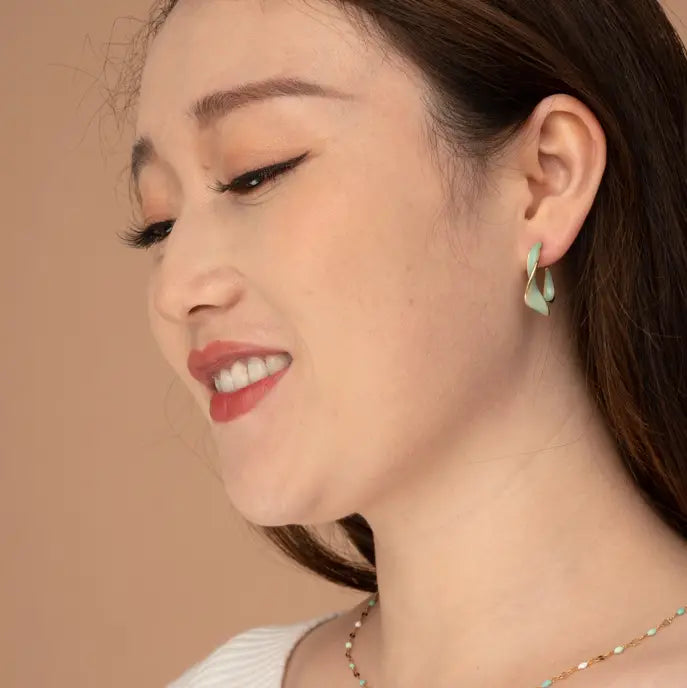 A close-up side profile of a woman wearing a light green crescent-shaped drop earring and a delicate multi-colored beaded necklace, set against a solid warm beige background.