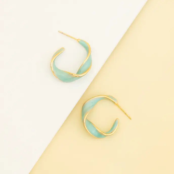 A pair of gold crescent-shaped hoop earrings with a light mint green enamel inlay, displayed on a diagonally split white and pale yellow background.