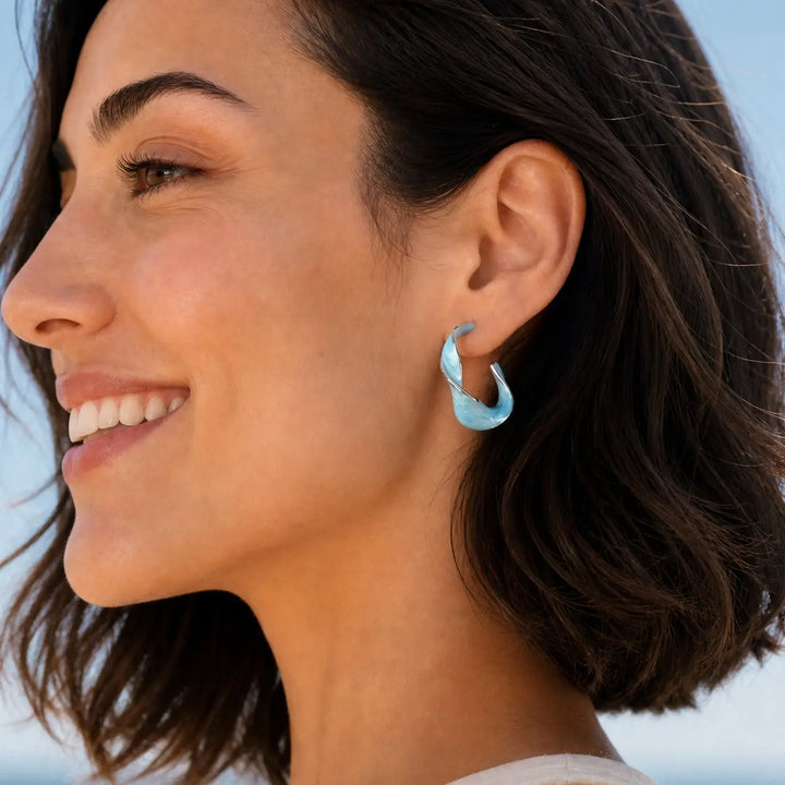 A close-up side profile of a woman wearing a light blue crescent-shaped hoop earring, set against a soft, out-of-focus light blue background.
